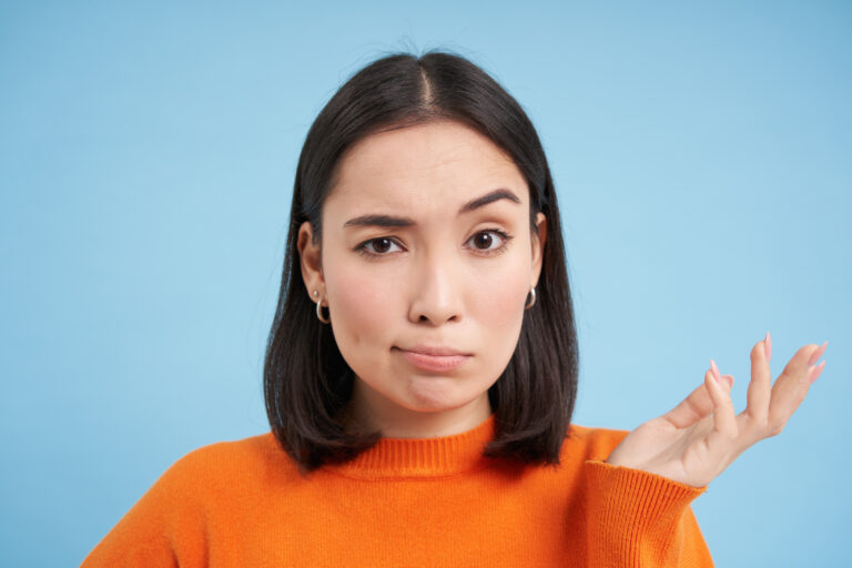 So what. Portrait of korean woman shrugs and raises arm up with skeptical, confused face expression, stands over blue background.